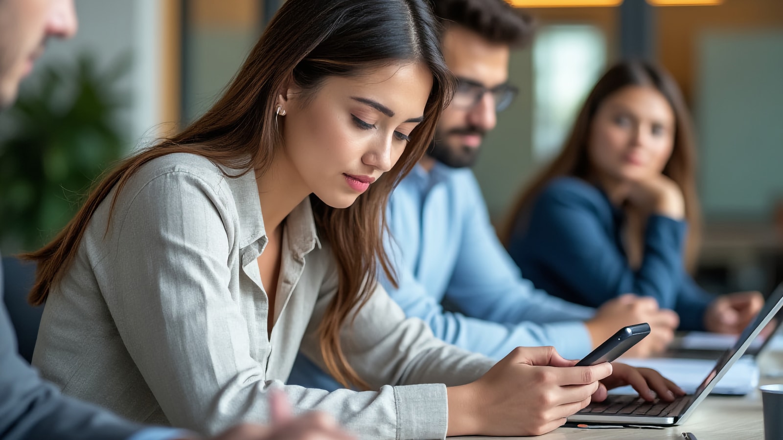 Mujer joven revisando su teléfono en una mesa de conferencias con colegas y computadoras portátiles de fondo