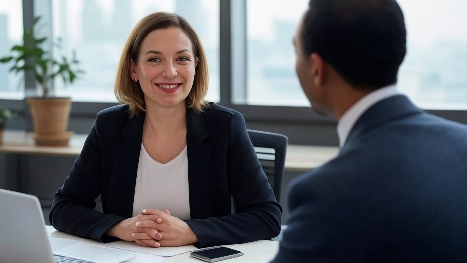 Mujer sonriente en blazer realizando una entrevista presencial en un escritorio de oficina con una computadora portátil