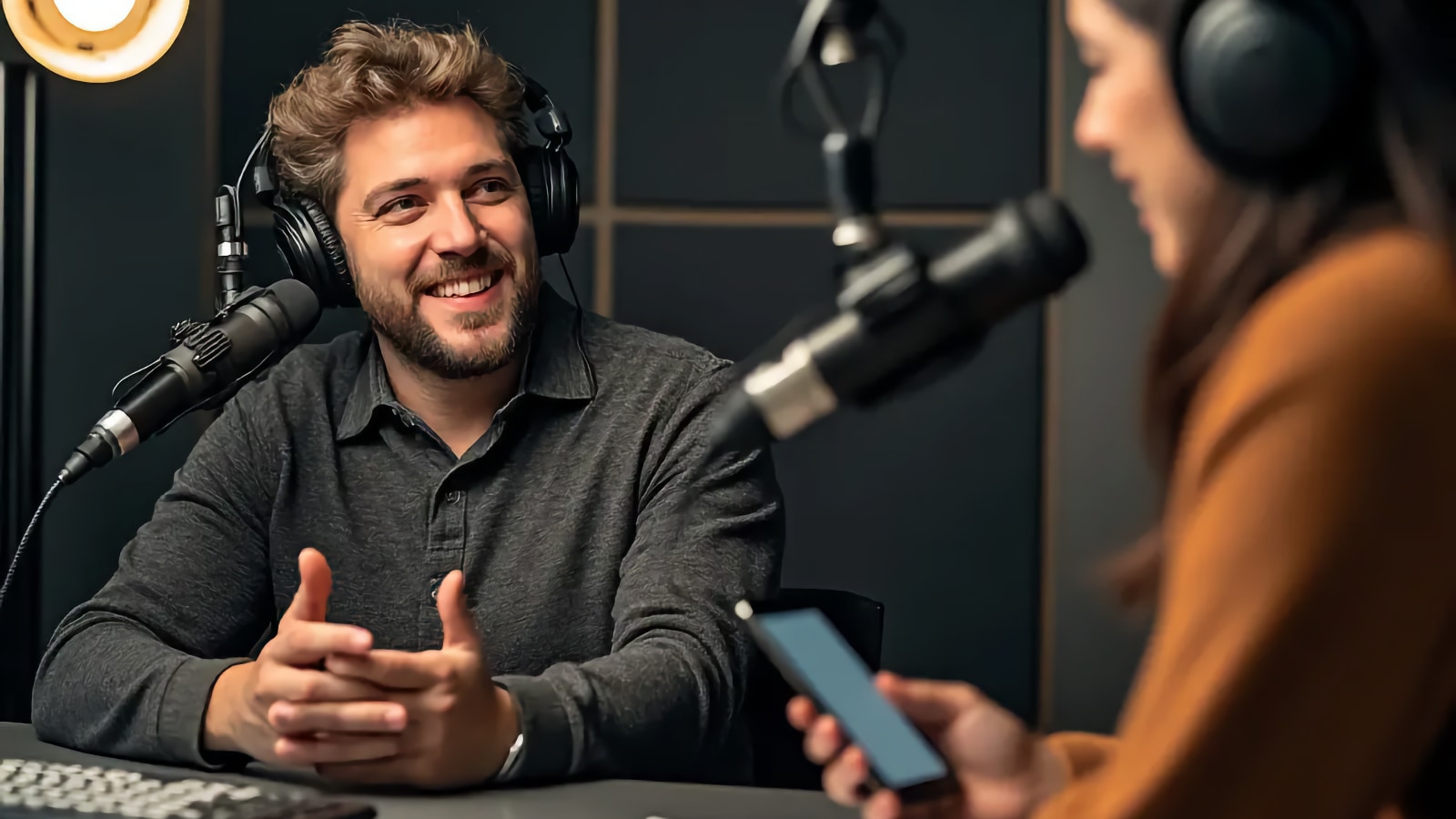 Hombre con auriculares hablando a un micrófono de estudio mientras una mujer sostiene un teléfono al otro lado del escritorio