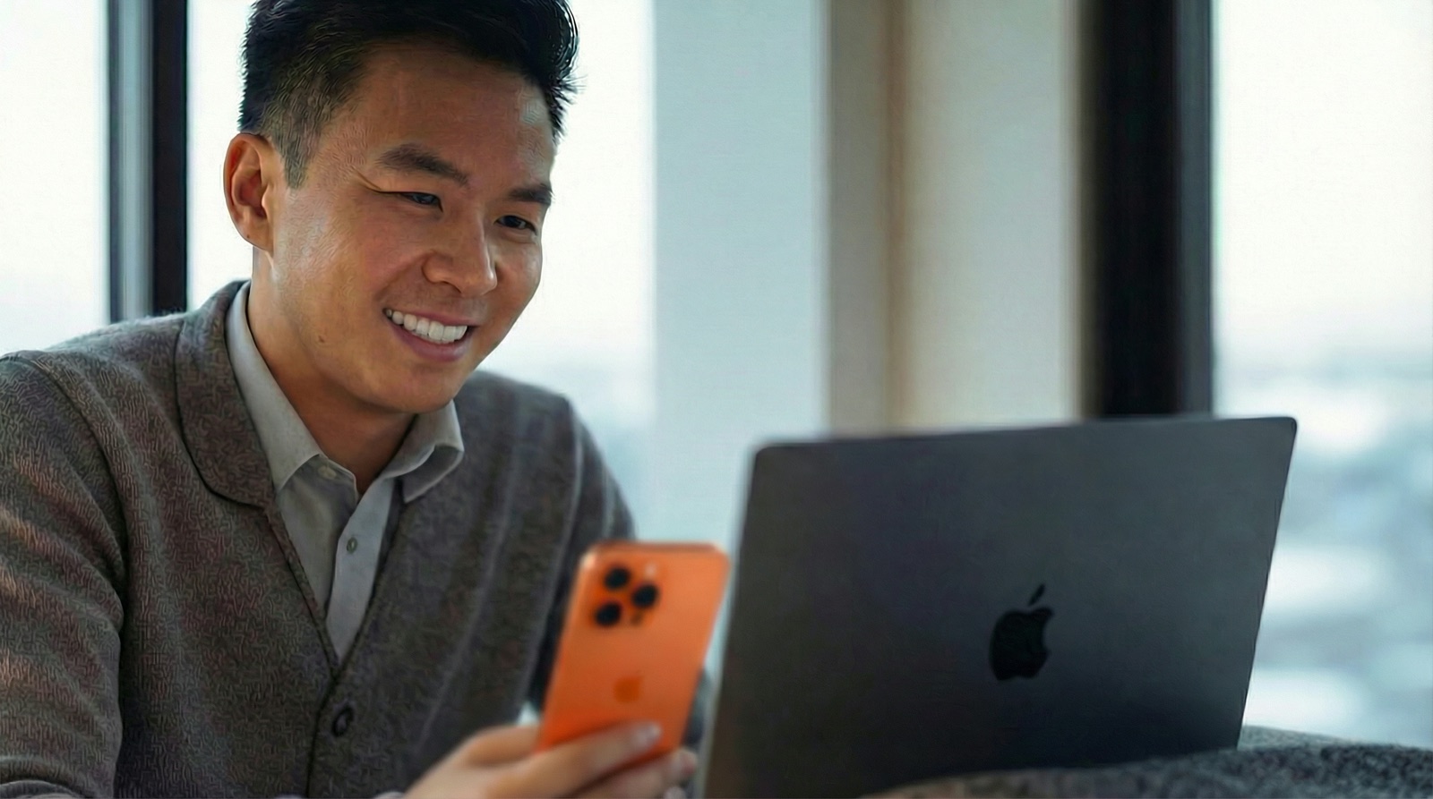 Hombre sonriente mirando un iPhone naranja junto a su laptop MacBook al lado de una ventana